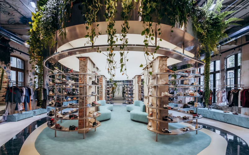 Interior da loja Mata Lab, um espaço moderno com conceito biofílico, onde estantes circulares exibem diversos modelos de calçados no centro do ambiente. Acima delas, uma estrutura metálica suspensa sustenta plantas pendentes e vegetação abundante. O espaço possui grandes janelas, iluminação natural, poltronas em tons claros e prateleiras com roupas nas laterais, criando uma atmosfera sofisticada e integrada à natureza.