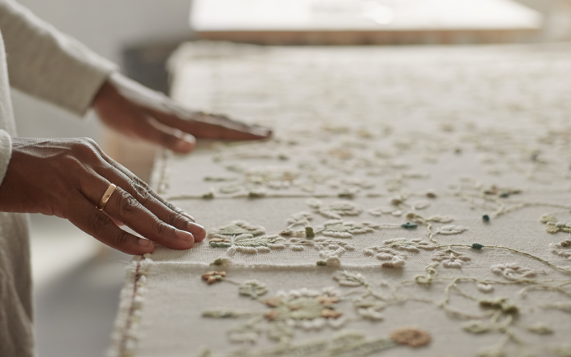 Mãos femininas tocando e ajustando um tecido bordado com detalhes florais, em um ambiente iluminado por luz natural, com foco na textura e no trabalho artesanal.